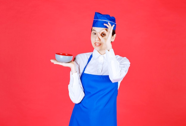 Cameriera della donna in uniforme che tiene una ciotola e che fa il gesto giusto vicino all'occhio sulla parete rossa.