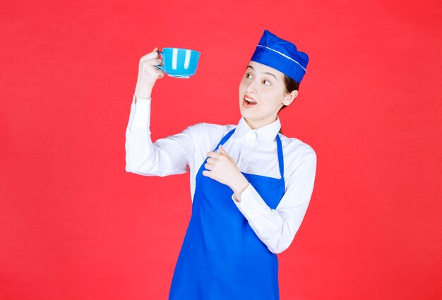 Cameriera donna in uniforme in piedi e con in mano una tazza blu sul muro rosso.