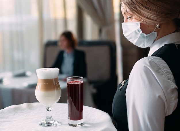 Cameriera femminile di aspetto europeo in una maschera medica serve caffè Latte.