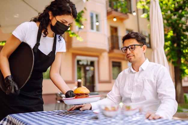 Cameriera in maschera protettiva che serve cibo delizioso per l'uomo in un accogliente ristorante.