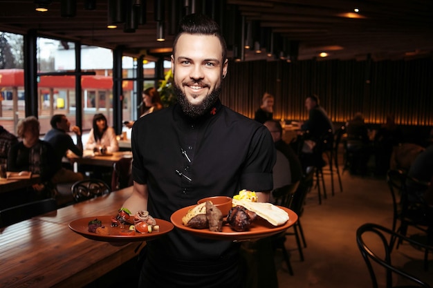 Cameriere barbuto allegro con kebab e insalata in un ristorante georgiano. Un cameriere esigente vestito di nero, con la barba e una porzione di kebab.