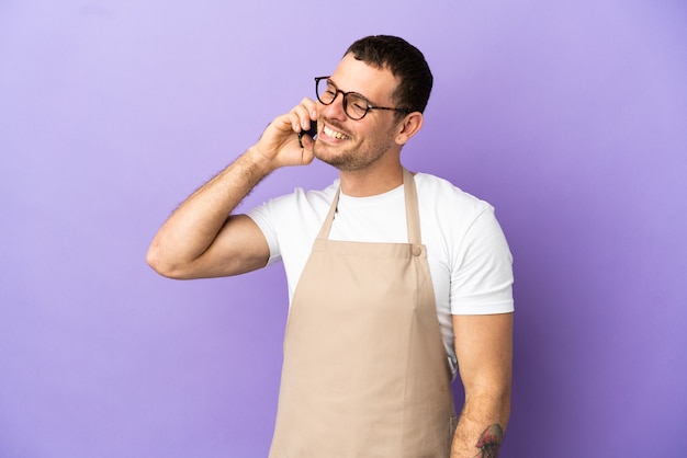 Cameriere brasiliano del ristorante su sfondo viola isolato che tiene una conversazione con il telefono cellulare