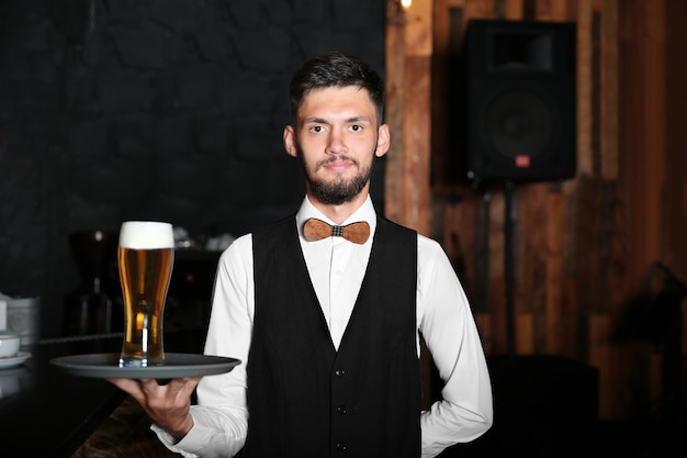 Cameriere che tiene il vassoio con un bicchiere di birra vicino al bancone del bar in legno