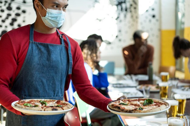 Cameriere con maschera protettiva che serve una deliziosa pizza agli amici nell'accogliente ristorante pizzeria Cameriere asiatico con due pizze Margherita in mano Ricetta originale della gustosa pizza margherita italiana