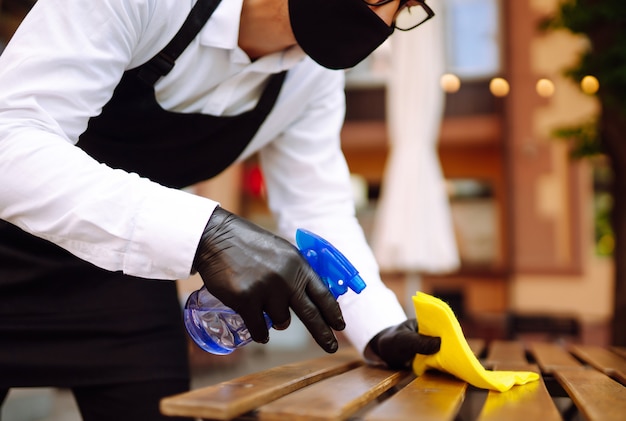 Cameriere con maschera protettiva e guanti che disinfettano il tavolo del ristorante per il prossimo cliente.