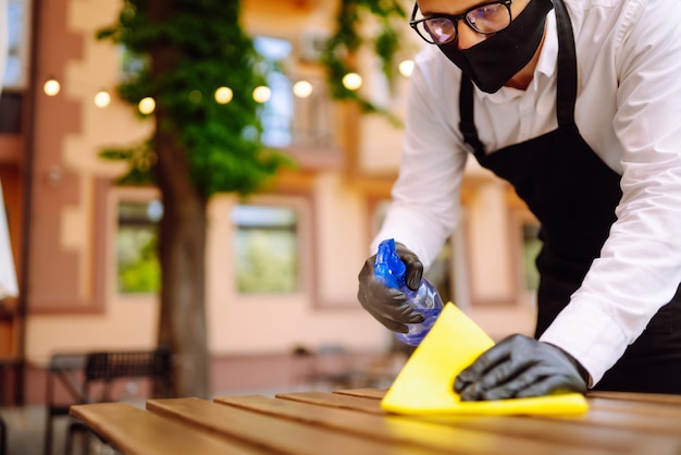 Cameriere con maschera protettiva e guanti che disinfettano il tavolo del ristorante per il prossimo cliente.