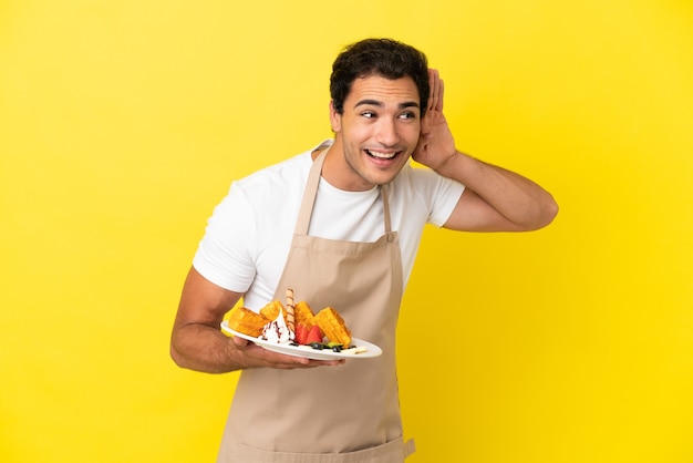 Cameriere del ristorante che tiene waffle su sfondo giallo isolato ascoltando qualcosa mettendo la mano sull'orecchio