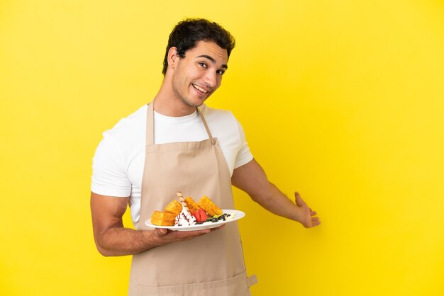 Cameriere del ristorante che tiene waffle su sfondo giallo isolato che allunga le mani di lato per invitare a venire