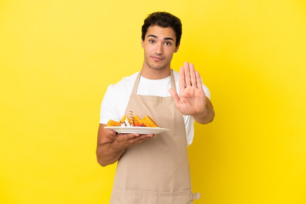 Cameriere del ristorante che tiene waffle su sfondo giallo isolato che fa un gesto di arresto