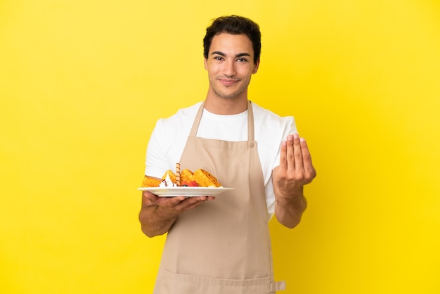 Cameriere del ristorante che tiene waffle su sfondo giallo isolato che invita a venire con la mano. Felice che tu sia venuto