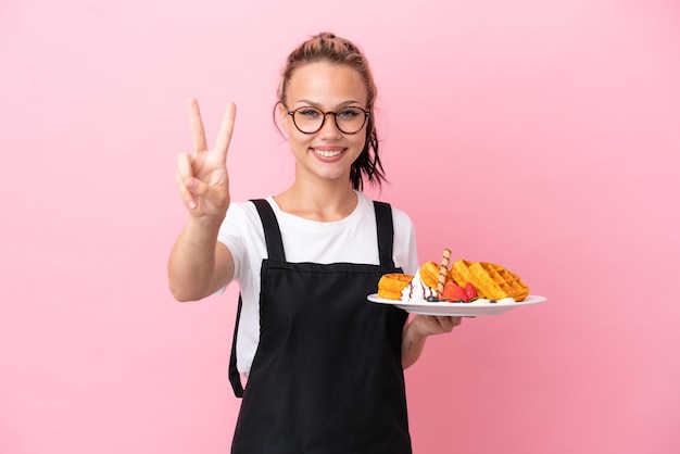 Cameriere del ristorante Ragazza russa con waffle isolati su sfondo rosa che sorride e mostra il segno della vittoria