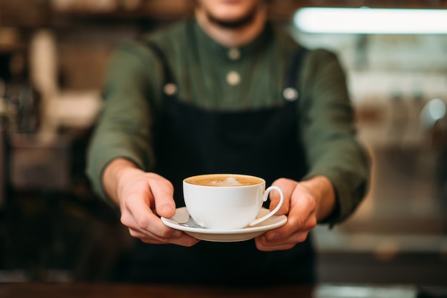 Cameriere in grembiule nero allunga una tazza di caffè con crema in mano.