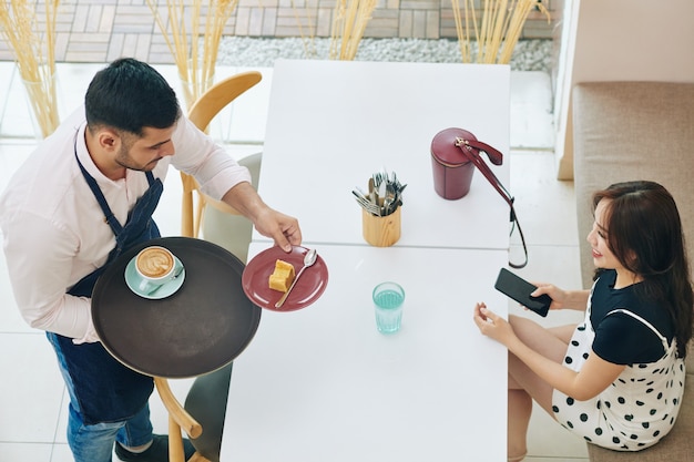 Cameriere portando un pezzo di torta deliziosa e una tazza di cappuccino alla bella giovane donna al tavolo del bar