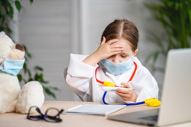 Camice e maschera d'uso sorridenti della piccola ragazza, orsacchiotto misurato di temperatura.