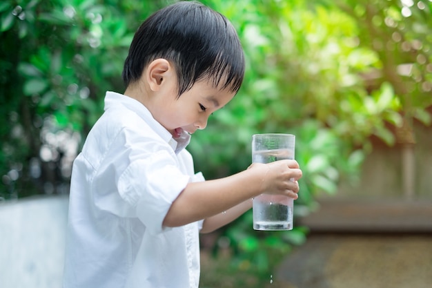 Camicia bianca di usura sveglia asiatica del ragazzo che beve acqua fredda da vetro nel fondo verde della natura