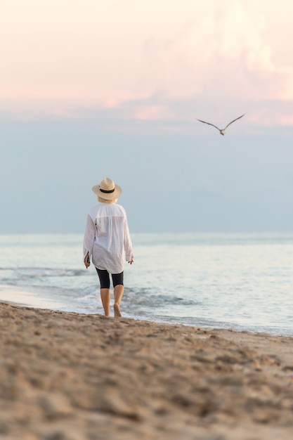 Camicia e cappello di paglia bianchi d'uso della donna che camminano sulla spiaggia al tramonto