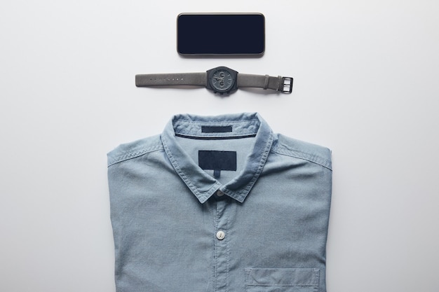 Camicia ripiegata, orologio da polso e telefono cellulare disposti in uno sfondo bianco isolato.