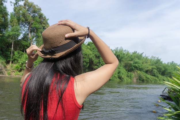 Camicia rossa della ragazza asiatica felice e cappello marrone sul fiume nel fondo della natura