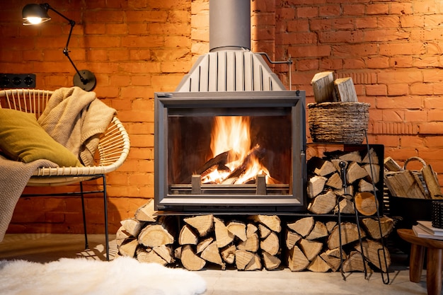 Camino accogliente con legna da ardere nell'interno della casa in stile loft con sfondo di muro di mattoni, fuoco ardente nel camino, intimità della casa in inverno