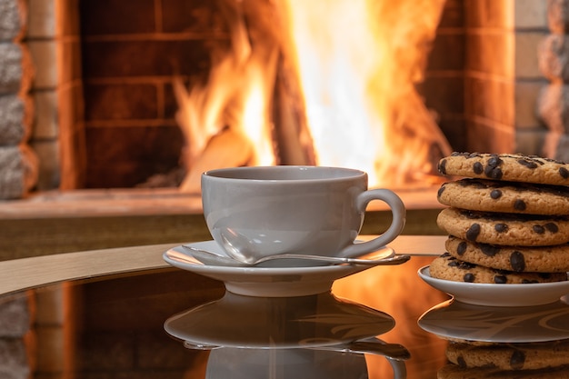 Camino accogliente, tazza di tè bianca e biscotti su un tavolo, riflesso di un fuoco su un tavolo di vetro.