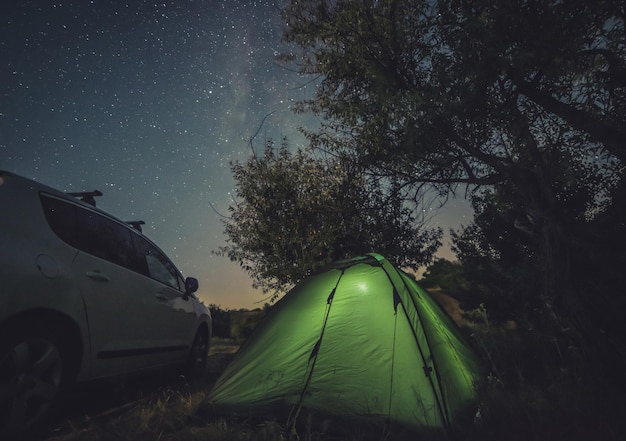 Campeggio con auto e tenda sotto l'auto turistica del cielo stellato e tenda verde sotto il cielo notturno luminoso