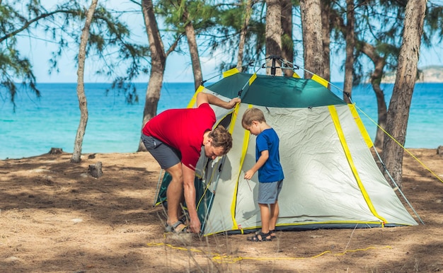 Campeggio persone all'aperto stile di vita turisti che organizzano il loro campeggio grigio verde nella foresta estiva vicino al mare di Lazur Ragazzo carino figlio aiuta suo padre a montare la tenda Educazione naturale dei bambini