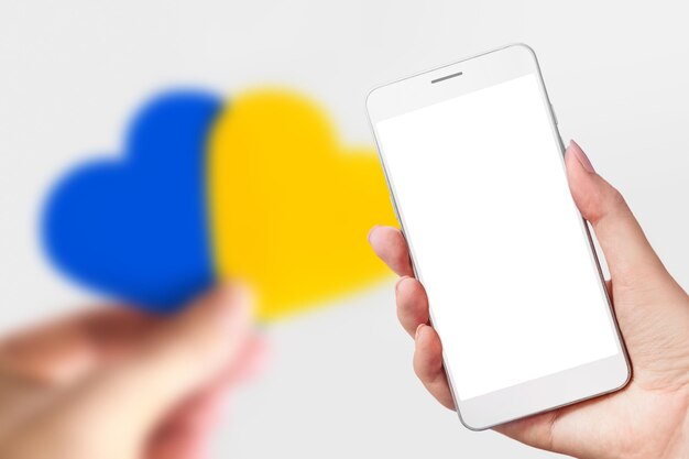 Canale di notizie ucraino La mano femminile tiene un telefono cellulare con uno schermo bianco vuoto e le mani femminili tengono un cuore con un'immagine della bandiera dell'Ucraina isolata su sfondo blu social network