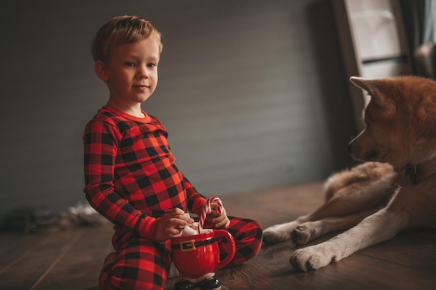 Candido autentico bambino felice in pigiama scozzese rosso abbraccia il cane con farfallino al lodge natale decorato