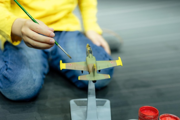 Candido bambino di sei anni che dipinge o colora o colora l'aeroplano modello con vernici a guazzo mentre si accovaccia sullo studio del pavimento illuminato da una luce bianca gialla brillante