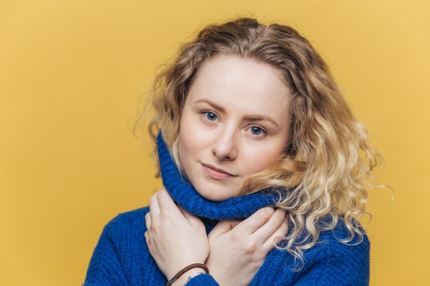 Candido colpo di seria adorabile donna con i capelli ricci indossa un caldo maglione blu si riscalda dopo una passeggiata all'aperto isolato su sfondo giallo studio La femmina attraente ha un'espressione sicura