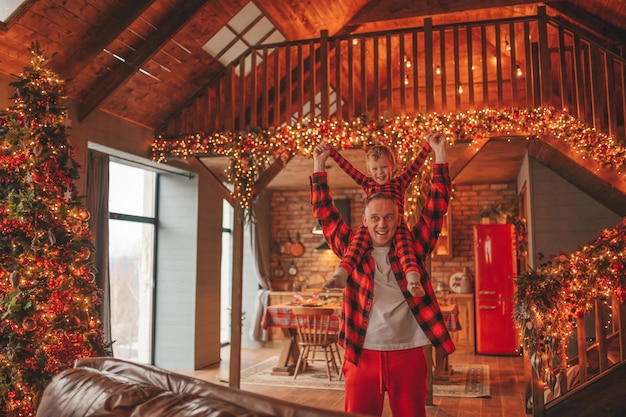 Candido padre e figlio felici e sinceri in pigiama scozzese rosso che scherzano al lodge di legno decorato per Natale