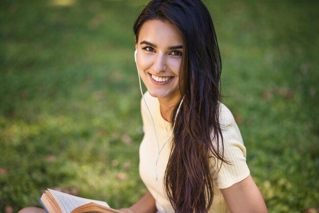 Candido ritratto di bruna caucasica bella giovane donna sorridente con gli auricolari che ascolta musica e legge un libro al parco Attraente donna felice che si rilassa nel parco durante il picnicCopia spazio