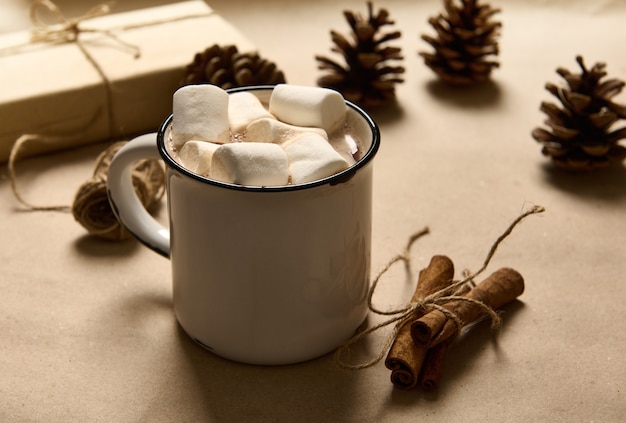 Cannella legate a prua da una corda accanto a una semplice vecchia tazza rustica di cioccolata calda e marshmallow dolci bianchi su uno sfondo di cartone con pigne e regalo di Natale in carta da regalo artigianale