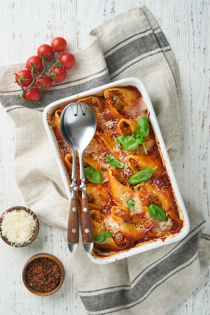 Cannelloni o conchiglioni Conchiglie di pasta ripiene al forno con sugo di carne bolognese pomodori basilico su tavola di legno bianca rustica Pasta al forno bolognese italiana tradizionale con parmigiano Cucina italiana