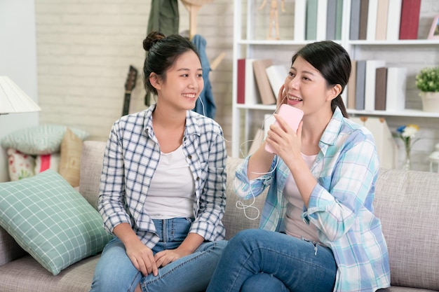 Cantiamo insieme. Belle amiche che ridono in abiti casual ascoltano musica dagli auricolari e tengono il telefono cellulare come microfono che canta sul divano. le donne allegre si rilassano in casa sorridendo