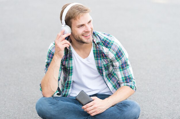 Canzone preferita. lo studente rilassa l'uso del telefono. ragazzo in chat all'aperto. l'uomo si siede a terra. studente spensierato in cuffia. tempo libero. formazione in linea. ascoltare musica. concetto di e-book. camicia a scacchi uomo.