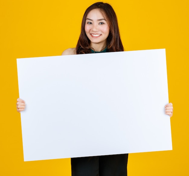 Capelli carini e piuttosto ricci Bruna femmina asiatica che tiene il bordo bianco bianco posa alla telecamera con un gioioso per scopi pubblicitari e banner, studio girato isolato su sfondo giallo brillante.