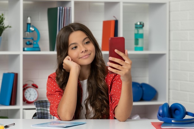 Capelli ricci lunghi del bambino felice che prendono la foto del selfie sullo smartphone nel vlogging dell'aula