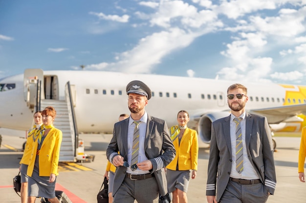 Capitani di aviazione maschi e assistenti di volo femminili che camminano vicino all'aereo passeggeri sotto il cielo nuvoloso in aeroporto