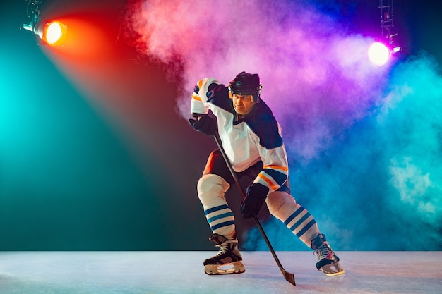 Capo. Giocatore di hockey maschio sul campo da ghiaccio e sfondo colorato al neon scuro con torce elettriche e fumo. Sportivo nella pratica dell'attrezzatura. Concetto di sport, stile di vita sano, movimento, benessere, azione.