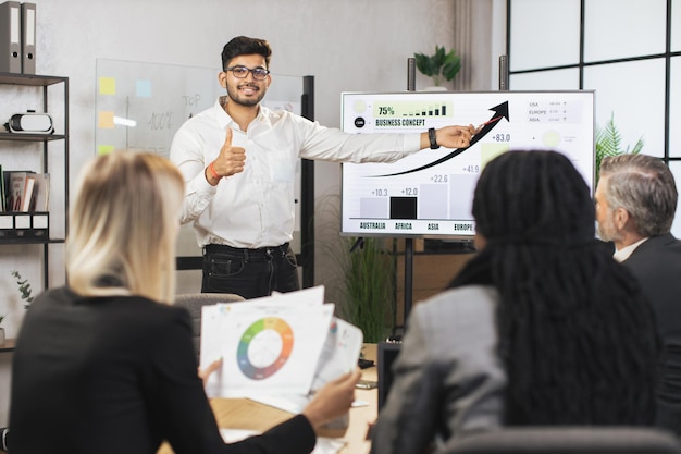 Capo indiano barbuto maschio di mezza età che mostra le infografiche di lavoro dell'azienda sullo schermo a parete digitale