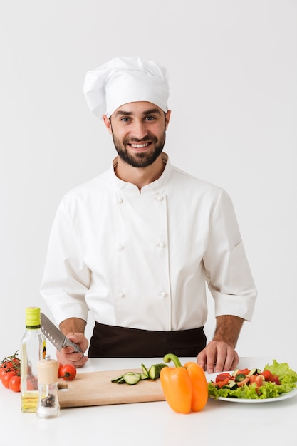 capo professionista in uniforme sorridente e cucinare insalata di verdure su tagliere di legno isolato su muro bianco