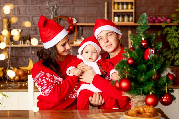 Capodanno o Natale, una giovane famiglia felice mamma, papà e bambino nella cucina buia della casa con un albero di Natale in maglioni rossi e cappelli di Babbo Natale sorridenti, abbracciati congratulandosi per la vacanza