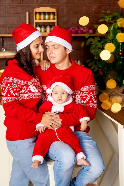 Capodanno o Natale, una giovane famiglia felice mamma, papà e bambino nella cucina buia della casa con un albero di Natale in maglioni rossi e cappelli di Babbo Natale sorridenti, abbracciati congratulandosi per la vacanza