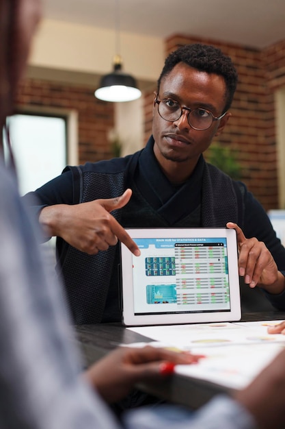 Capogruppo dell'agenzia di ricerca e marketing che indica i dati delle statistiche aziendali sul tablet. Uomo d'affari afroamericano che informa il collega dell'ufficio sulla necessità del taglio del budget.