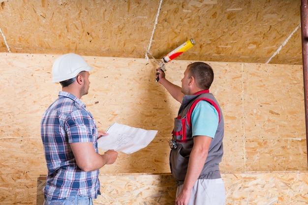 Caposquadra del sito in costruzione che indossa elmetto bianco che tiene i piani e osservando il lavoratore applicando il calafataggio al soffitto in legno non finito