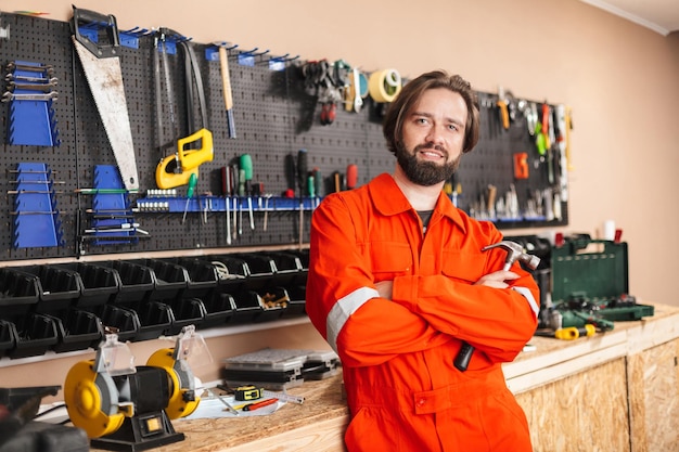 Caposquadra sorridente in abiti da lavoro arancioni che tiene il martello in mano guardando felicemente in camera con supporto di diversi strumenti sullo sfondo in officina