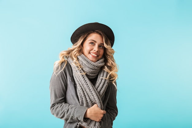 Cappello da portare della ragazza graziosa e una sciarpa che sta isolata sopra la parete blu