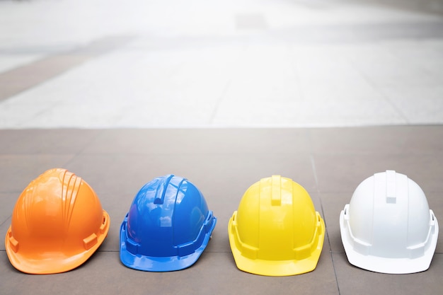 Cappello del casco di usura di sicurezza duro arancione, giallo, blu e bianco nel progetto al cantiere edile sul pavimento di cemento sulla città