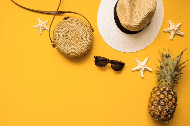 Cappello di paglia, borsa di bambù, occhiali da sole, ananas e stelle marine sopra il giallo